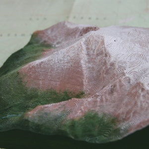 K&ouml;nnte beinhalten: Ein 3D-gedrucktes Modell einer Bergkette mit gr&uuml;nen und wei&szlig;en Details. Das Modell besteht aus einem strukturierten Material und hat ein realistisches Aussehen.
