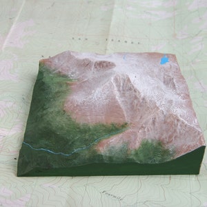 K&ouml;nnte beinhalten: Ein 3D-Modell einer Bergkette mit schneebedeckten Gipfeln und einem gr&uuml;nen Tal. Das Modell besteht aus einem strukturierten Material und ist in Braun-, Gr&uuml;n- und Wei&szlig;t&ouml;nen bemalt.