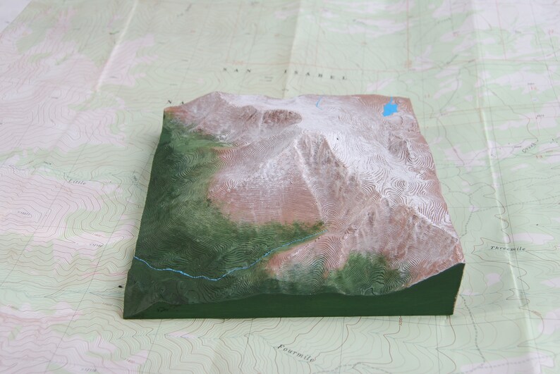 K&ouml;nnte beinhalten: Ein 3D-Modell einer Bergkette mit schneebedeckten Gipfeln und einem gr&uuml;nen Tal. Das Modell besteht aus einem strukturierten Material und ist in Braun-, Gr&uuml;n- und Wei&szlig;t&ouml;nen bemalt.