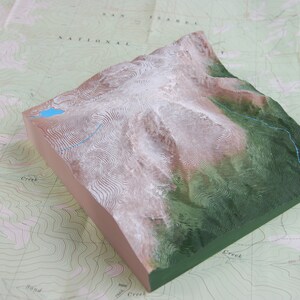 K&ouml;nnte beinhalten: Ein 3D-Modell einer Bergkette mit einem blauen See an der Spitze. Das Modell ist aus hellem braunen Material gefertigt und hat eine gr&uuml;ne Basis. Das Modell steht auf einer Karte mit H&ouml;henlinien.