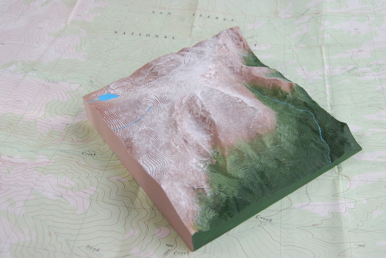 K&ouml;nnte beinhalten: Ein 3D-Modell einer Bergkette mit einem blauen See an der Spitze. Das Modell ist aus hellem braunen Material gefertigt und hat eine gr&uuml;ne Basis. Das Modell steht auf einer Karte mit H&ouml;henlinien.