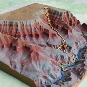 May include: A detailed, three-dimensional model of a canyon landscape. The model features reddish-brown cliffs, a winding golden path, and a blue river. The terrain is mounted on a brown base, suggesting a topographical representation.