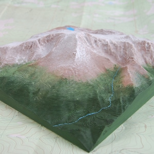 K&ouml;nnte beinhalten: Ein 3D-gedrucktes Modell einer Bergkette mit gr&uuml;nem Sockel und braunen und wei&szlig;en Gipfeln. Das Modell befindet sich auf einer Karte mit H&ouml;henlinien.