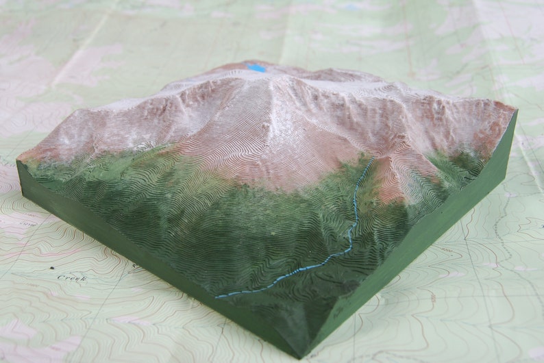 K&ouml;nnte beinhalten: Ein 3D-gedrucktes Modell einer Bergkette mit gr&uuml;nem Sockel und braunen und wei&szlig;en Gipfeln. Das Modell befindet sich auf einer Karte mit H&ouml;henlinien.