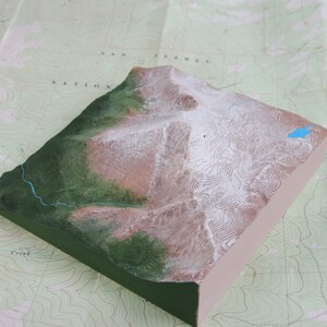 K&ouml;nnte beinhalten: Ein 3D-Modell einer Bergkette mit schneebedeckten Gipfeln und einem kleinen blauen See. Das Modell besteht aus einem hellbraunen Material mit gr&uuml;nen und wei&szlig;en Details. Das Modell steht auf einer Karte mit H&ouml;henlinien.