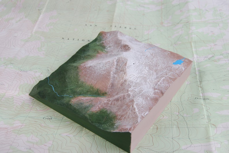 K&ouml;nnte beinhalten: Ein 3D-Modell einer Bergkette mit schneebedeckten Gipfeln und einem kleinen blauen See. Das Modell besteht aus einem hellbraunen Material mit gr&uuml;nen und wei&szlig;en Details. Das Modell steht auf einer Karte mit H&ouml;henlinien.