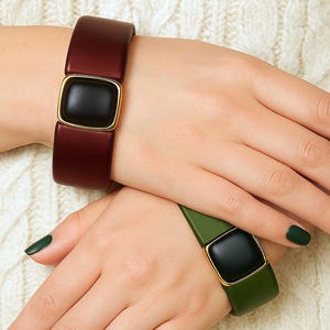 Könnte beinhalten: Zwei breite, rechteckige Armbänder mit einem quadratischen, schwarzen Mittelteil. Ein Armband ist weinrot mit einem goldfarbenen Rahmen um das schwarze Quadrat. Das andere Armband ist olivgrün mit einem goldfarbenen Rahmen um das schwarze Quadrat. Die Armbänder befinden sich auf einem weißen Zopfstrickpullover.
