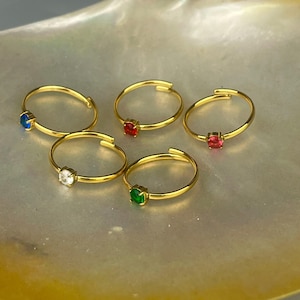 Peut inclure: Cinq bagues réglables de couleur or, chacune ornée d'une pierre précieuse de couleur différente : bleu, rouge, blanc, vert et rose. Les bagues sont présentées sur une surface nacrée, mettant en valeur les couleurs vives et le design des bijoux.