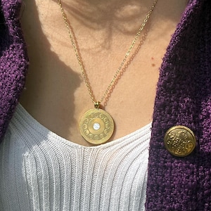 Könnte beinhalten: Eine goldene Halskette mit einem runden Anhänger, der ein Mondphasen-Design zeigt. Der Anhänger hat eine weiße Mitte und ist von einem Kreis aus kleineren Kreisen umgeben.