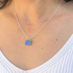 Könnte beinhalten: Eine silberne Halskette mit einem quadratischen Anhänger mit einem blauen Edelstein. Der Anhänger hängt an einer feinen Kette. Die Halskette wird über einem weißen Rippen-Top getragen.