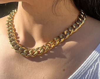 May include: A thick gold chain necklace with a curb link design. The necklace is worn by a person with dark hair and a white shirt.