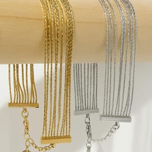 May include: Close-up of two multi-strand bracelets. One is gold-toned, the other is silver-toned. Each bracelet features multiple thin strands attached to a rectangular bar and a chain clasp. The bracelets are displayed on a wooden dowel.