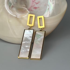 Peut inclure: Boucles d'oreilles rectangulaires dorées avec incrustation de nacre. Les boucles d'oreilles ont un dessus rectangulaire et un bas long et rectangulaire. Le fond présente un coquillage.