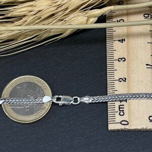 Op de afbeelding: Een zilveren slangenketting met een karabijn sluiting. De ketting ligt op een zwarte ondergrond naast een liniaal en een euro munt.