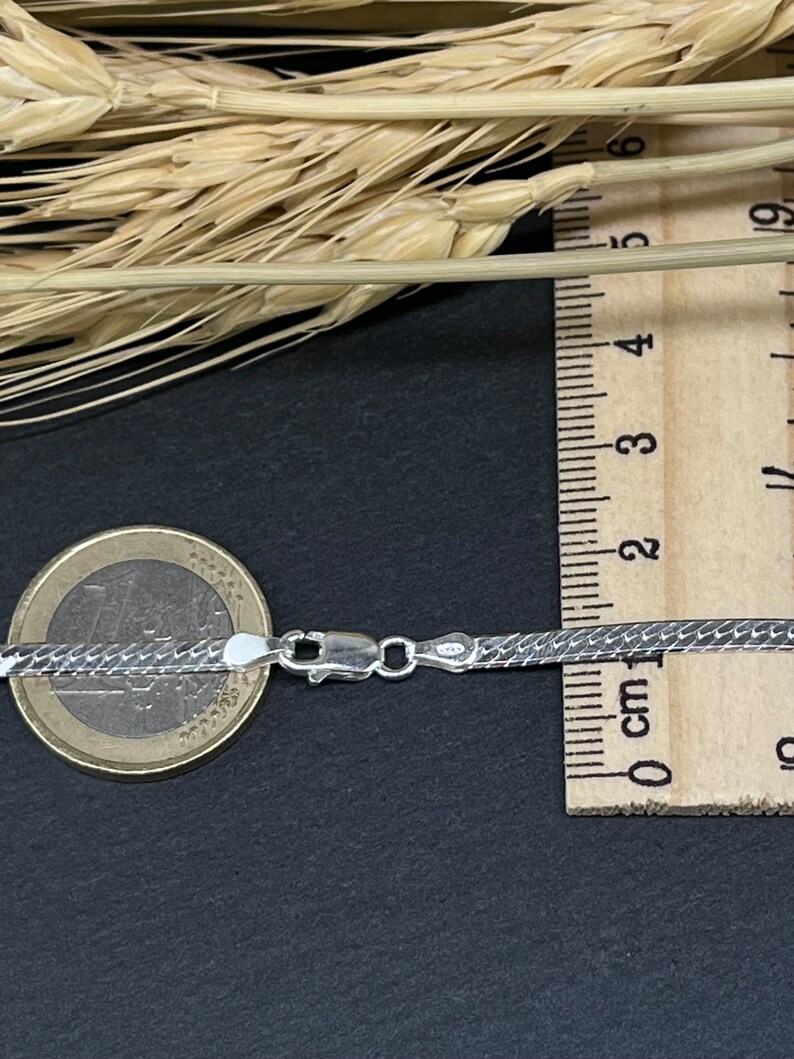 Op de afbeelding: Een zilveren slangenketting met een karabijn sluiting. De ketting ligt op een zwarte ondergrond naast een liniaal en een euro munt.