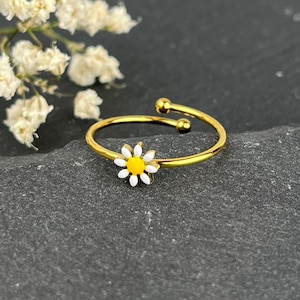 Könnte beinhalten: Ein verstellbarer Goldring mit einer weißen Gänseblümchen-Blume mit gelber Mitte.