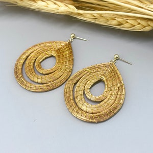 Könnte beinhalten: Ein Paar geflochtene Strohohrringe mit einem Spiraldesign. Die Ohrringe sind hellbraun und haben einen goldfarbenen Metallhaken.