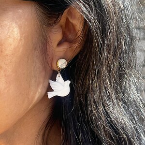 Puede incluir: Pendientes de cerámica blanca en forma de paloma con un poste dorado y un dije redondo blanco. Los pendientes son usados por una persona con cabello largo y oscuro.
