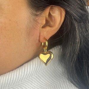 Puede incluir: Pendientes de aro dorados con un gran dije en forma de corazón brillante que cuelga de cada aro.