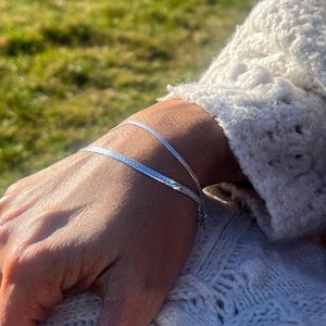 Peut inclure: Un bracelet argenté à chevrons est porté au poignet. Le bracelet fin et plat reflète la lumière. La personne porte un pull blanc en tricot texturé. L'arrière-plan est une vue floue d'herbe verte.