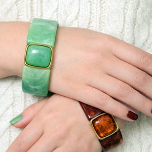 Könnte beinhalten: Zwei klobige Armbänder mit goldfarbenen quadratischen Akzenten. Eines ist marmoriert grün, das andere schildpattbraun. Die Armbänder werden an Handgelenken vor einem weißen, strukturierten Hintergrund getragen. Die Nägel sind grün und braun lackiert.