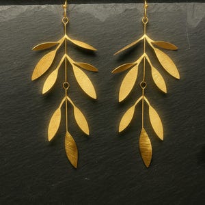Peut inclure: Une paire de boucles d'oreilles dorées en forme de feuilles. Chaque boucle d'oreille présente une série de feuilles stylisées suspendues à un crochet. Les feuilles sont disposées en cascade, avec une surface texturée. Les boucles d'oreilles sont présentées sur un fond sombre.
