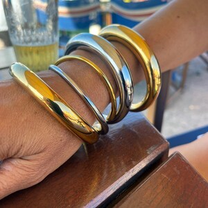 Peut inclure: Plusieurs bracelets dorés et argentés sont empilés sur un poignet. Les bracelets ont une surface polie et réfléchissante et varient en largeur. Les bracelets sont présentés sur le bras d'une personne, avec une table en bois au premier plan.