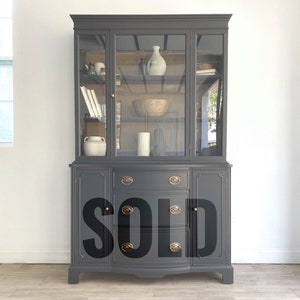 May include: A dark gray painted vintage hutch with glass doors and brass hardware. The hutch has a top cabinet with shelves and a lower cabinet with drawers. The hutch is sold.
