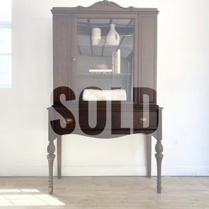 May include: A dark wood hutch with glass doors and a single drawer. The hutch has turned legs and brass hardware. The hutch is sold.