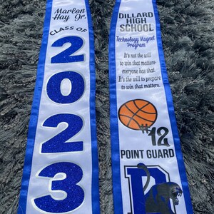 May include: White and blue graduation sash with the text "Marlon Hay Jr. Class of 2023" on one side and "Dillard High School Technology Magnet Program" on the other side. The sash also features a basketball, the number 12, the words "Point Guard", and a black panther.