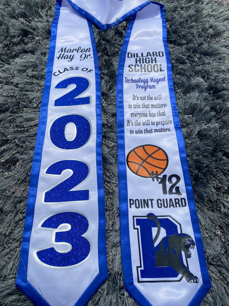 May include: White and blue graduation sash with the text "Marlon Hay Jr. Class of 2023" on one side and "Dillard High School Technology Magnet Program" on the other side. The sash also features a basketball, the number 12, the words "Point Guard", and a black panther.
