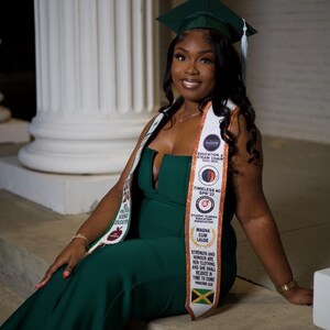 May include: A woman wearing a green graduation gown and cap, a white sash with gold lettering, and a gold necklace. The sash has the text "Education & Steam Chair 2022-2023", "Timeless 40 SPR 22", "Student Florida Education Association", "Magna Cum Laude", and "Strength and Honour are her clothing and she shall rejoice in time to come".