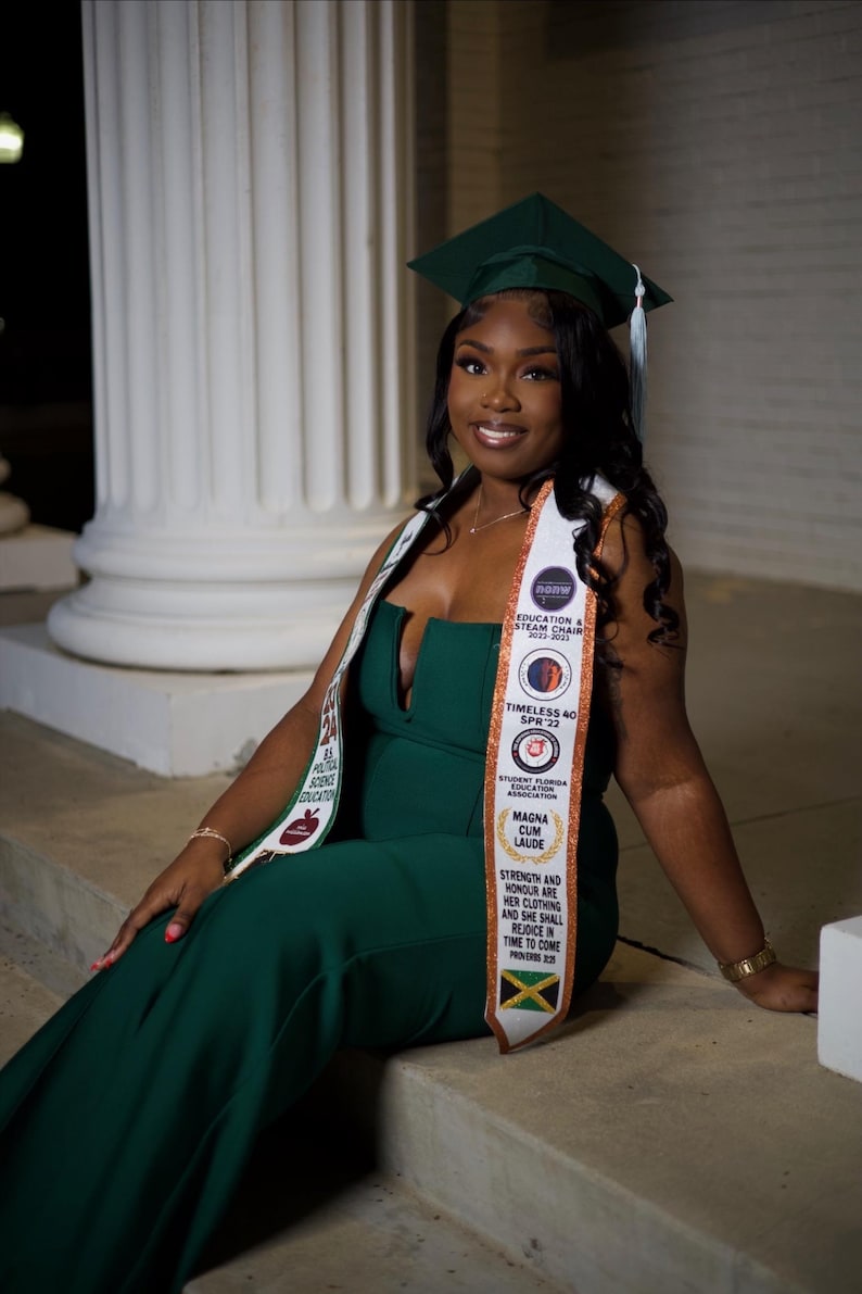May include: A woman wearing a green graduation gown and cap, a white sash with gold lettering, and a gold necklace. The sash has the text "Education & Steam Chair 2022-2023", "Timeless 40 SPR 22", "Student Florida Education Association", "Magna Cum Laude", and "Strength and Honour are her clothing and she shall rejoice in time to come".