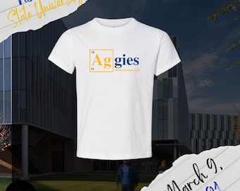Pode incluir: Camiseta branca com a palavra "Aggies" em azul e amarelo, com "North Carolina A&T" abaixo. A camiseta também tem o texto "NC A&T State University" e "March 9, 1891". A camiseta é exibida em frente a um edifício.