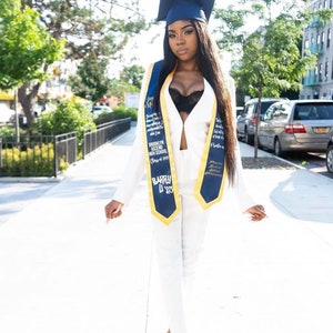 May include: A woman in a white pantsuit and black bra, wearing a blue and gold graduation sash, poses for a photo. She is wearing a black cap and gown and has long black hair. The sash has the text "Brooklyn Ascend High School Class of 2023" and "Barry L'23".