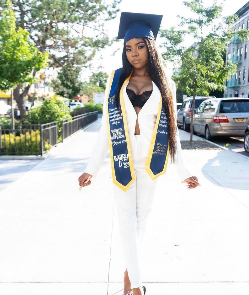 May include: A woman in a white pantsuit and black bra, wearing a blue and gold graduation sash, poses for a photo. She is wearing a black cap and gown and has long black hair. The sash has the text "Brooklyn Ascend High School Class of 2023" and "Barry L'23".