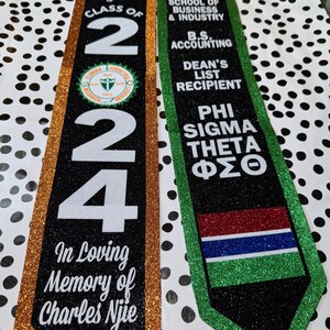 May include: Two graduation sashes, one gold and one green, with black glitter. The gold sash has the text "Sirra Njanko Njie Class of 2024" and "In Loving Memory of Charles Njie". The green sash has the text "Florida Agricultural & Mechanical University School of Business & Industry B.S. Accounting Dean's List Recipient Phi Sigma Theta &Phi;&Sigma;&Theta;".