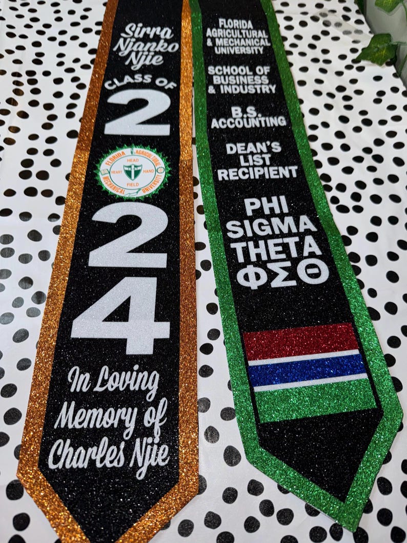 May include: Two graduation sashes, one gold and one green, with black glitter. The gold sash has the text "Sirra Njanko Njie Class of 2024" and "In Loving Memory of Charles Njie". The green sash has the text "Florida Agricultural & Mechanical University School of Business & Industry B.S. Accounting Dean's List Recipient Phi Sigma Theta &Phi;&Sigma;&Theta;".