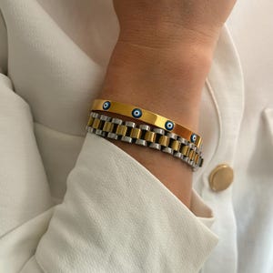 May include: Two bracelets are shown. The top bracelet is gold-colored with three blue eye-shaped charms. The bottom bracelet is silver and gold-colored, with a watch-band style design. The bracelets are worn on a wrist, with a white shirt in the background.