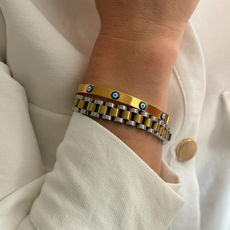 May include: Two bracelets are shown. The top bracelet is gold-colored with three blue eye-shaped charms. The bottom bracelet is silver and gold-colored, with a watch-band style design. The bracelets are worn on a wrist, with a white shirt in the background.