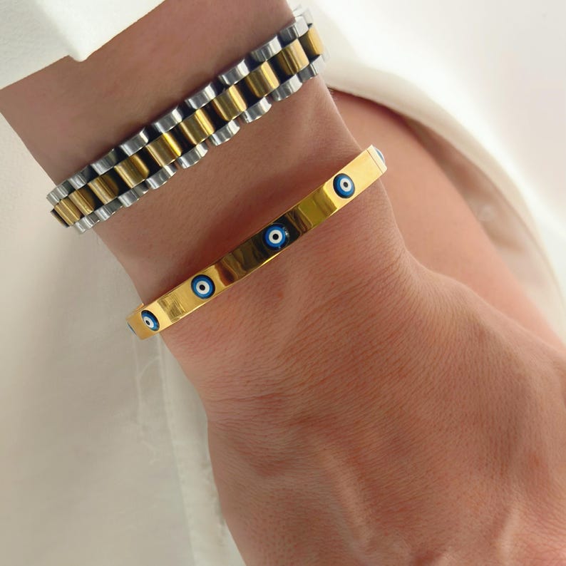 May include: Two bracelets on a wrist. One is a gold and silver watch-style bracelet. The other is a gold bangle with blue and white evil eye designs. The bracelets are worn on a person's wrist.