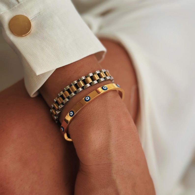 May include: A gold-toned bangle bracelet with blue eye designs and a silver and gold-toned watch band bracelet. The bracelets are stacked on a wrist, with a white shirt and gold cufflink in the background.