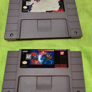 K&ouml;nnte beinhalten: Zwei graue Super Nintendo-Spielmodule. Eines hat ein besch&auml;digtes Etikett, das andere zeigt ein farbenfrohes Bild mit dem Text "THE FIGHTING EDITION" und "SUPER NINTENDO". Die Module liegen auf einer gr&uuml;nen Oberfl&auml;che.