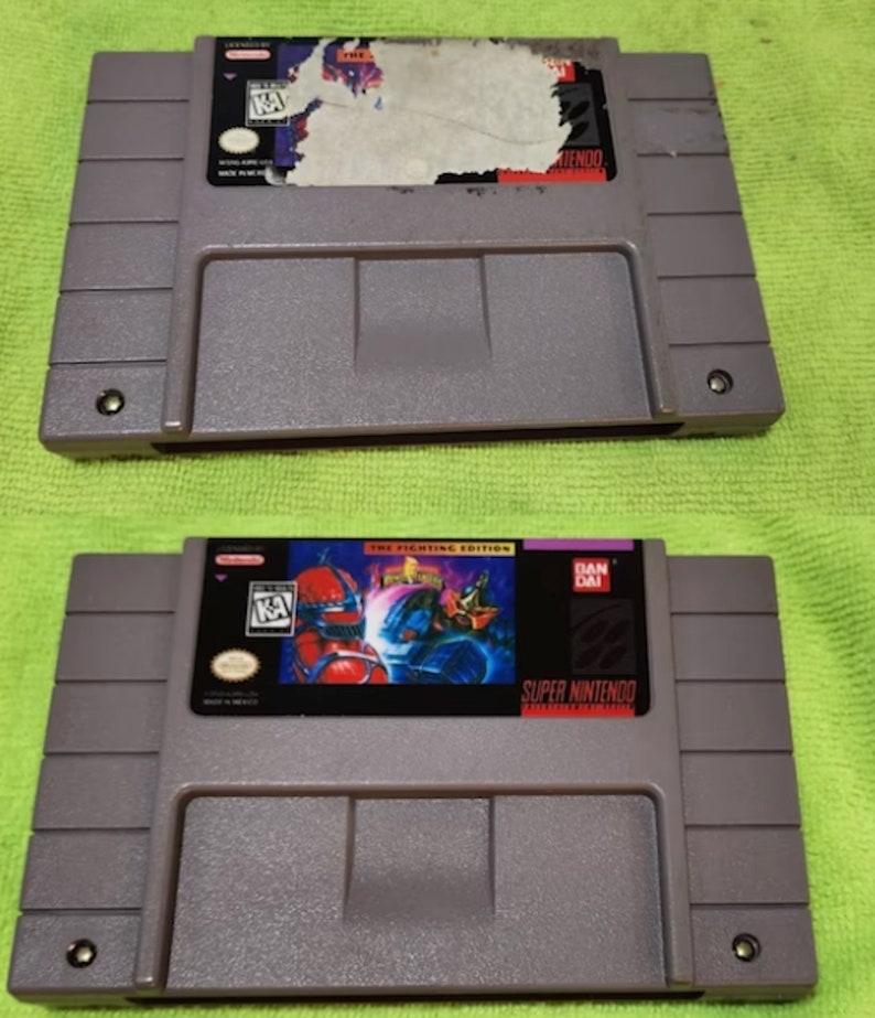 K&ouml;nnte beinhalten: Zwei graue Super Nintendo-Spielmodule. Eines hat ein besch&auml;digtes Etikett, das andere zeigt ein farbenfrohes Bild mit dem Text "THE FIGHTING EDITION" und "SUPER NINTENDO". Die Module liegen auf einer gr&uuml;nen Oberfl&auml;che.