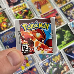 K&ouml;nnte beinhalten: Eine Nahaufnahme eines Pokemon-Videospiel-Kartuschen-Etiketts. Das Etikett ist silberfarben mit einem roten, orangefarbenen und gelben Bild eines Pokemon-Charakters. Das Etikett enth&auml;lt auch den Text "Pokemon", "Nintendo", "Jedermann" und "ESRB".