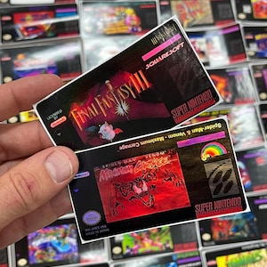 K&ouml;nnte beinhalten: Zwei Etiketten von Super Nintendo-Spielkassetten werden hochgehalten, mit vielen weiteren im Hintergrund. Das obere Etikett ist f&uuml;r Final Fantasy III, und das untere Etikett ist f&uuml;r Spider-Man & Venom: Maximum Carnage.