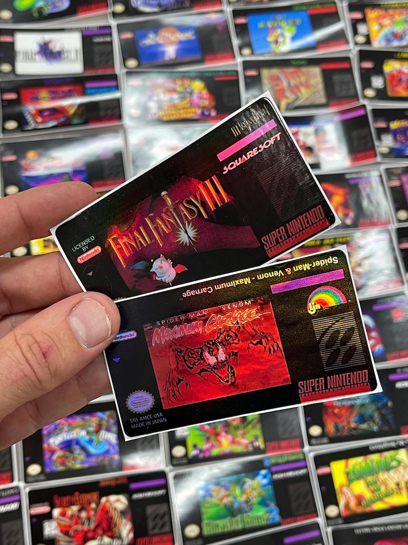 K&ouml;nnte beinhalten: Zwei Etiketten von Super Nintendo-Spielkassetten werden hochgehalten, mit vielen weiteren im Hintergrund. Das obere Etikett ist f&uuml;r Final Fantasy III, und das untere Etikett ist f&uuml;r Spider-Man & Venom: Maximum Carnage.
