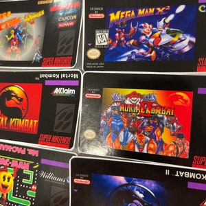 K&ouml;nnte beinhalten: Eine Sammlung von Super Nintendo-Spielkassetten mit Titeln wie "Mortal Kombat", "Mega Man X2" und "Ms. Pac-Man". Die Spielboxen haben farbenfrohe Grafiken und das Super Nintendo-Logo. Die Spiele sind von Nintendo lizenziert.