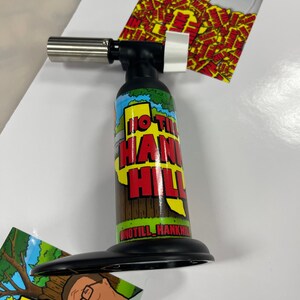 May include: A black and silver butane torch with a red, yellow, and green graphic of a cartoon character and the text "HO TILL HANK HILL".