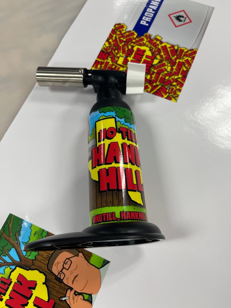 May include: A black and silver butane torch with a red, yellow, and green graphic of a cartoon character and the text "HO TILL HANK HILL".